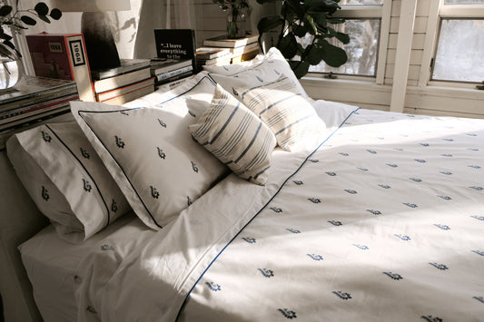 Blue Paisley bedsheet set on a sunlit bed with striped cushions, navy paisley motifs and navy piping on white percale
