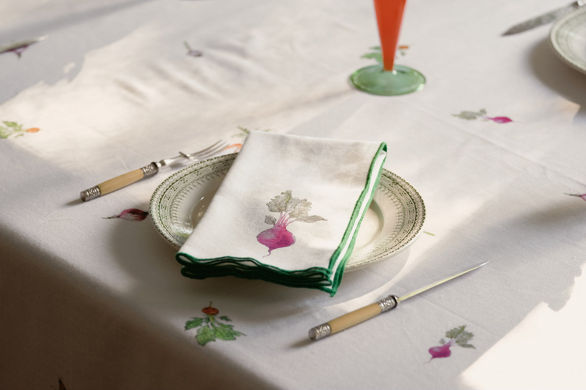 Red Beets print napkin with a green overlock hem folded on a plate on the Root Vegetable Tablecloth