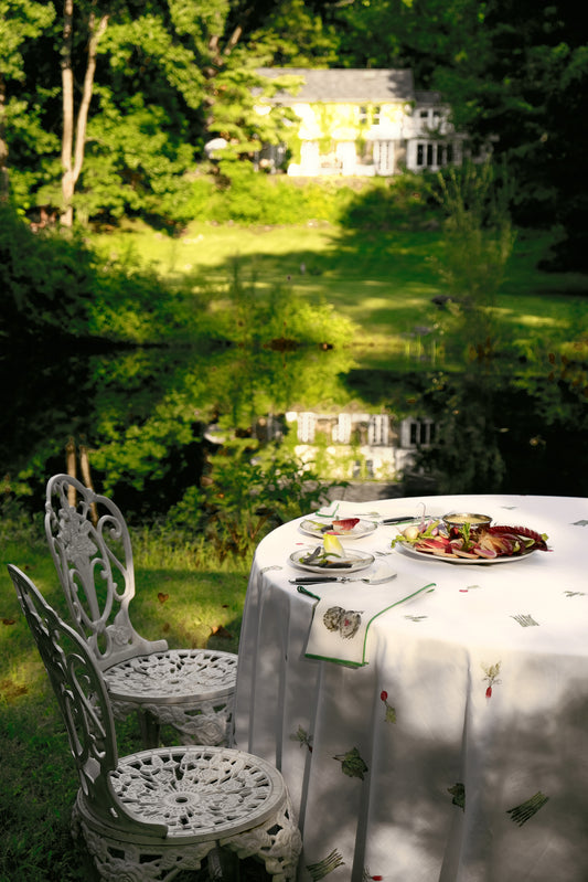 Spring Vegetable Tablecloth on a round table on a garden lawn, beside white iron chairs, with a pond and house in the distance