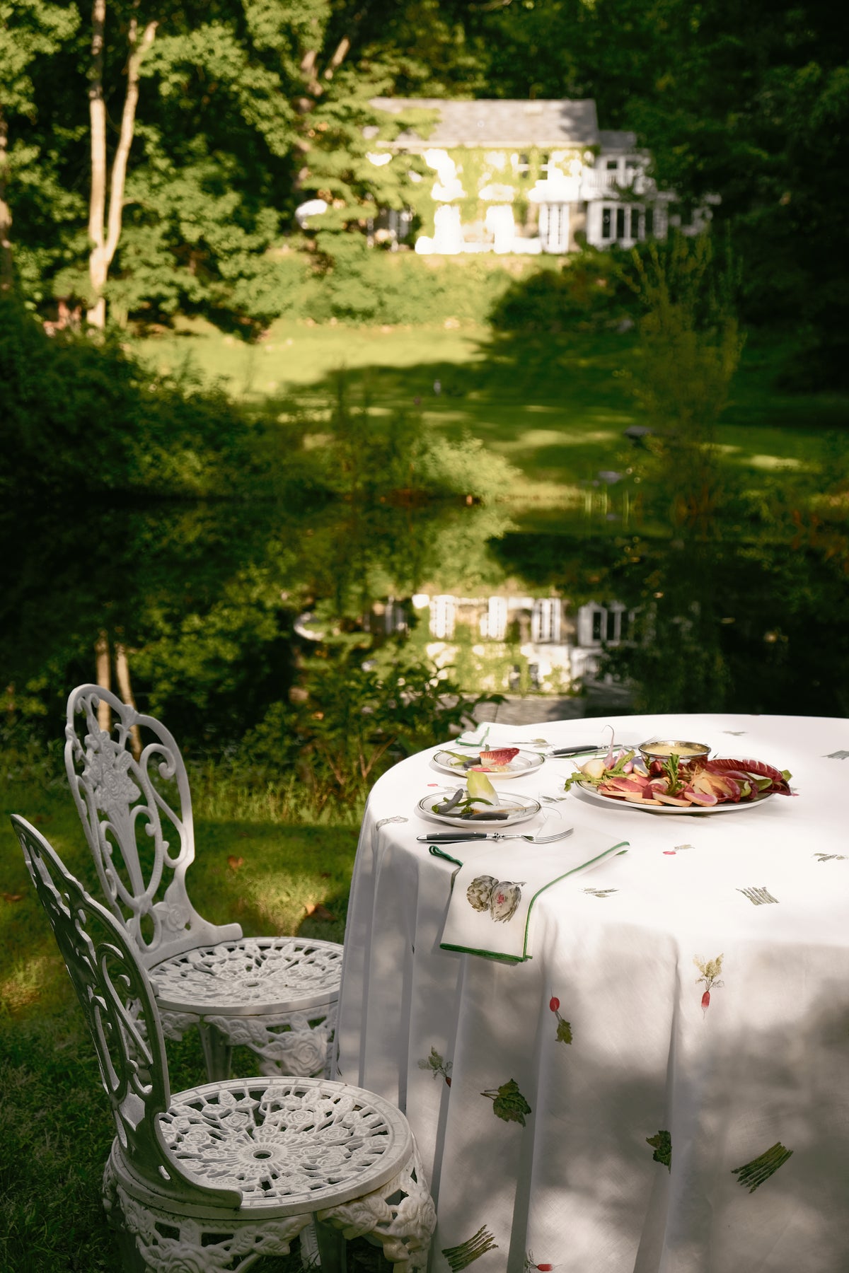 Spring Vegetable Tablecloth on a round table on a garden lawn, beside white iron chairs, with a pond and house in the distance