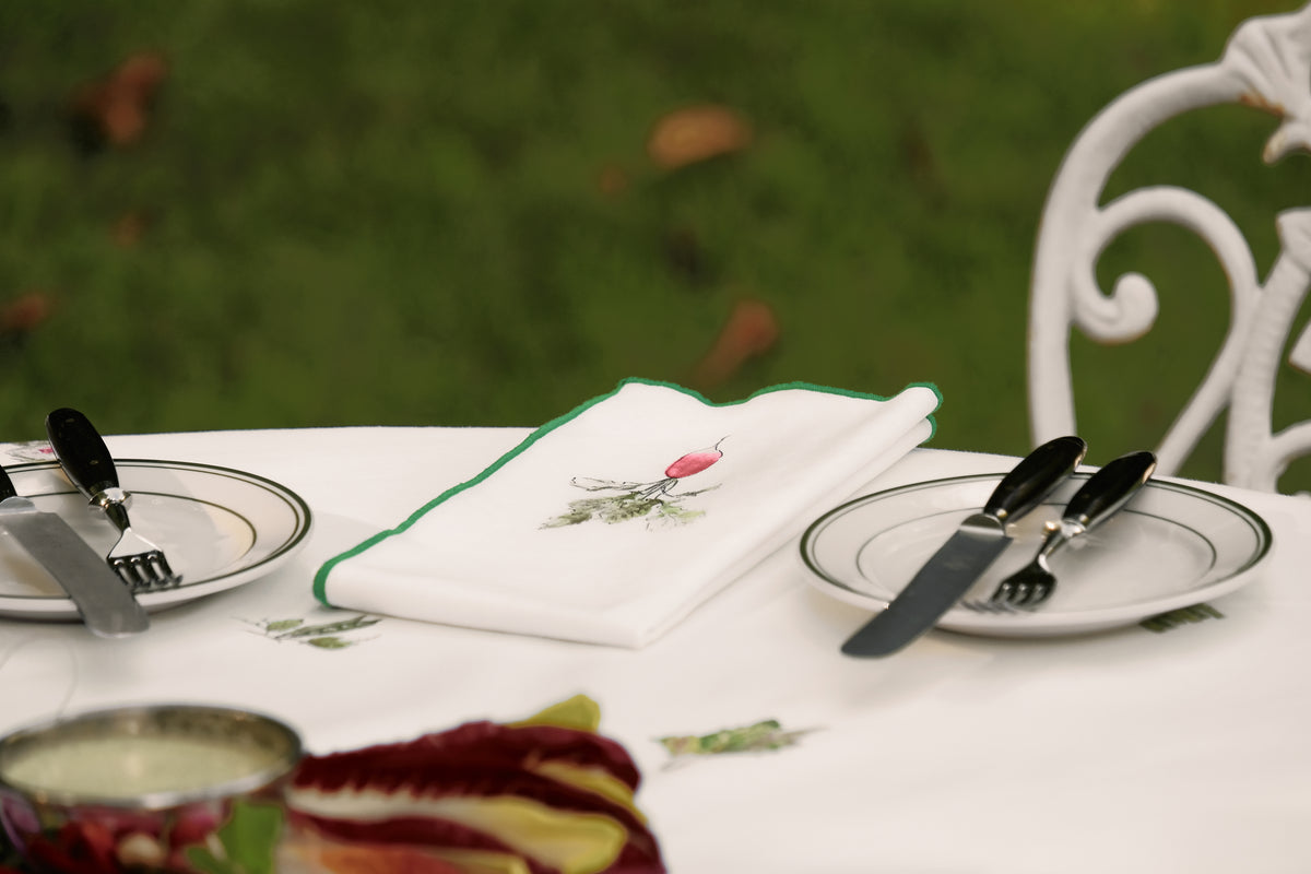 Radish print napkin with a green overlock hem, folded between two place settings on the Spring Vegetable Tablecloth