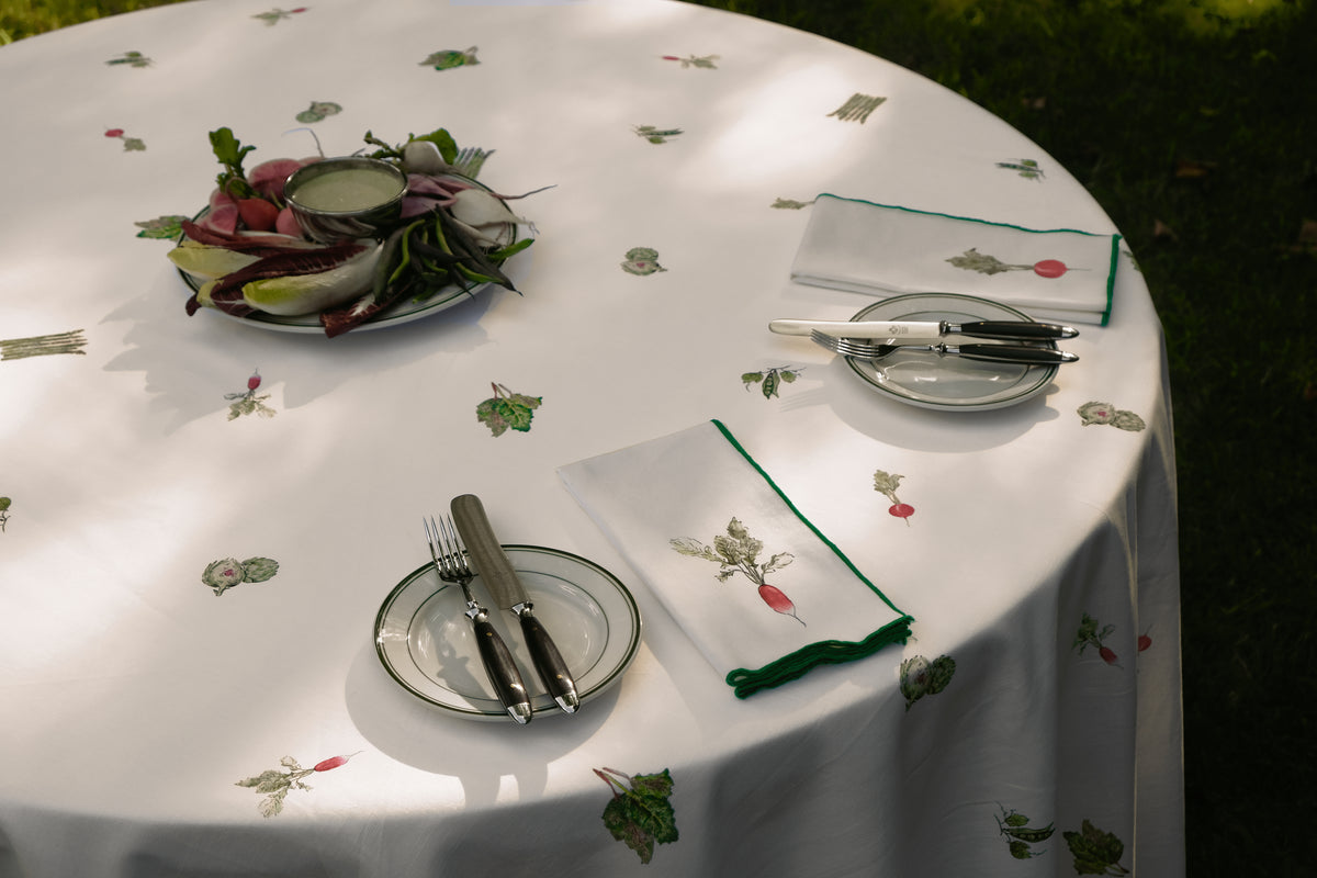 Spring Vegetable Tablecloth on a round table outdoors, set with radish print napkins, green-rimmed plates and a platter of spring vegetables